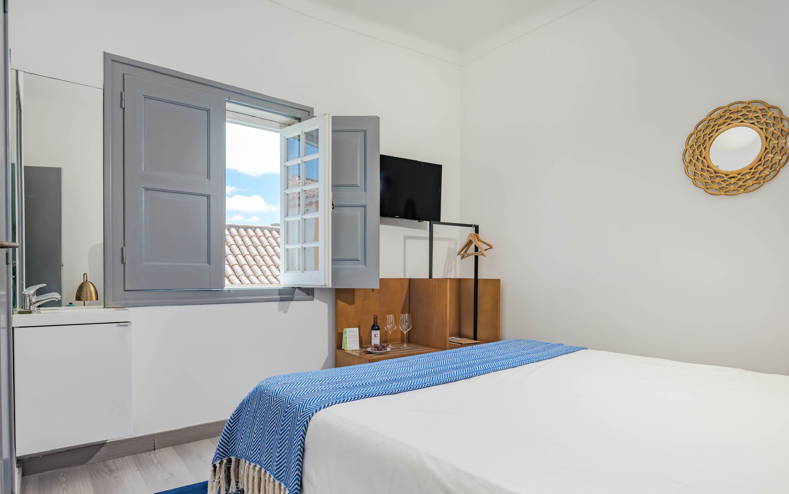 A hotel room with a bed, a TV mounted on the wall, a wooden dresser, and a window with open shutters.