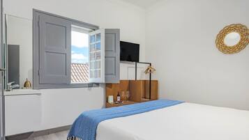 A hotel room with a bed, a TV mounted on the wall, a wooden dresser, and a window with open shutters.