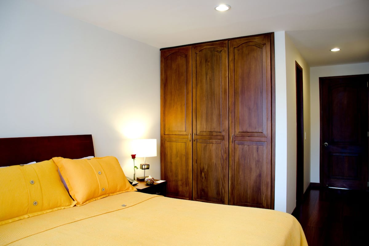 Une chambre d’hôtel avec une grande armoire en bois, un lit avec une literie jaune et une table de chevet avec une lampe et un vase.