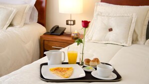 A hotel room with a neatly made bed, a bedside table with a lamp, a tray with breakfast items, and a rose on the bed. - Hotel Portofino (Bogotá)
