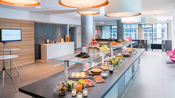 A modern hotel lobby with a reception desk, a buffet with various food items, and a large window providing a view of the city. - Barceló Hamburg (Hamburg)