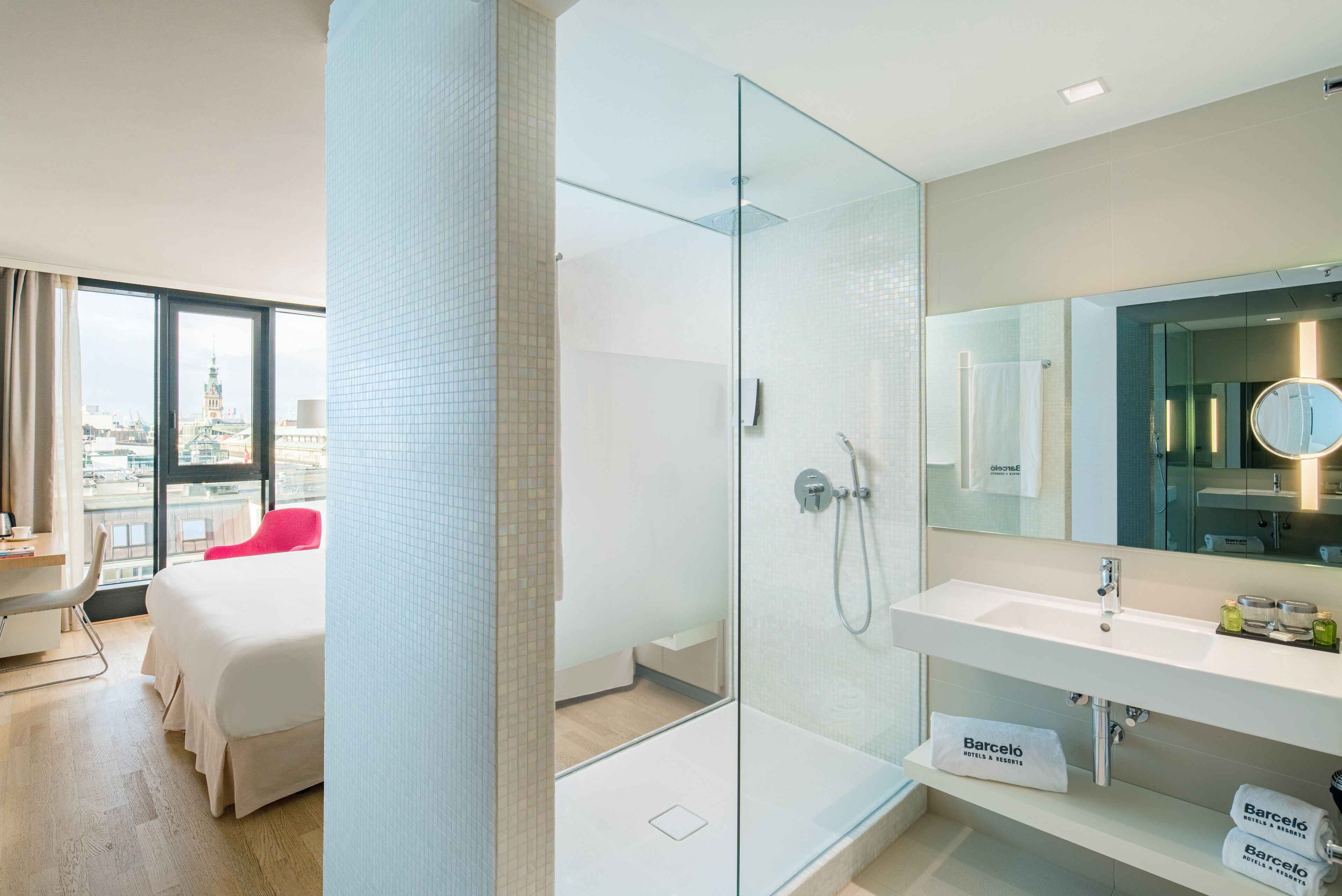 Une chambre d’hôtel moderne dotée d’une douche en verre, d’un lit blanc, d’un fauteuil rouge, d’une grande fenêtre donnant sur la ville et d’un mini-bar.