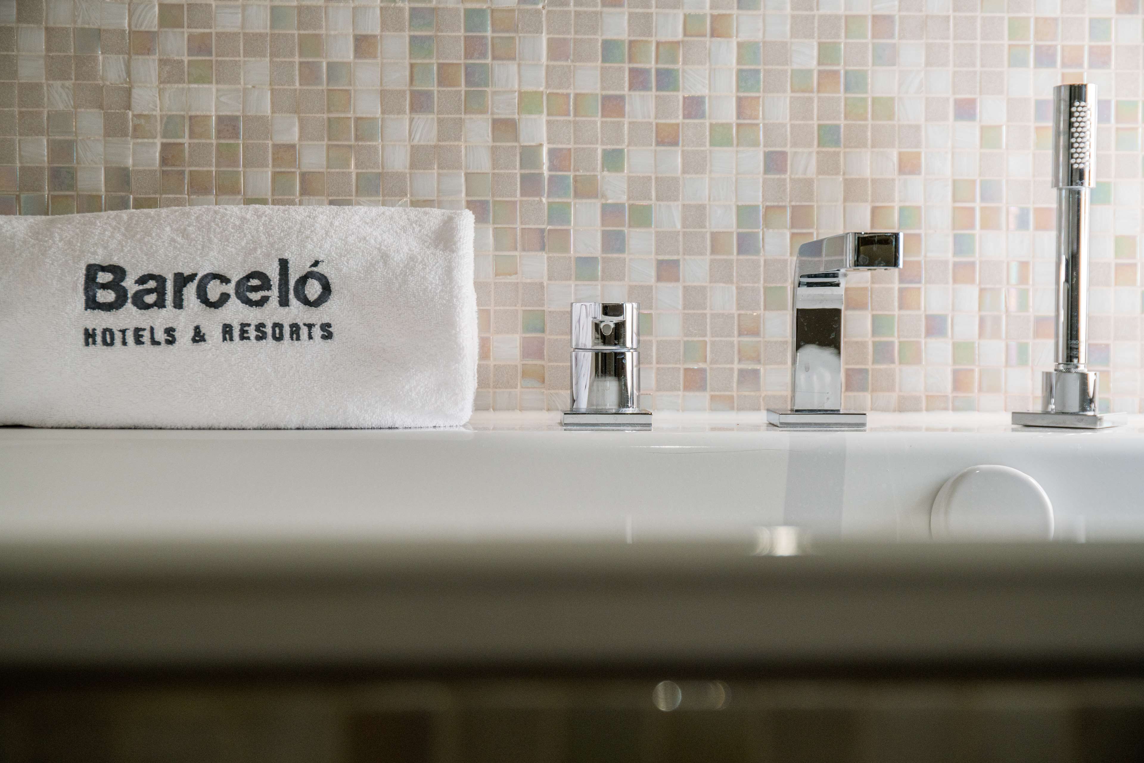 A bathroom with a tiled backsplash, a towel with "Barceló Hotels & Resorts" text, and two chrome faucets.