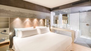 A modern hotel room with a large bed, a built-in vanity with a mirror, and a shower area. - Barceló Hamburg (Hamburg)
