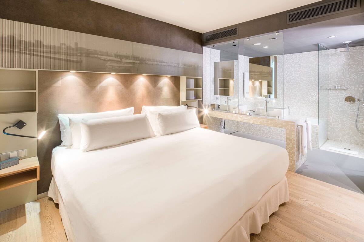 A modern hotel room with a large bed, a built-in vanity with a mirror, and a shower area.