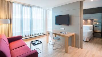 Ein modernes Hotelzimmer mit Flachbildfernseher, Esstisch, rotem Sofa und einem Bett mit weißer Daunendecke.