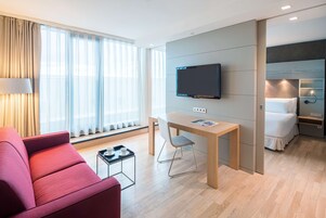 Ein modernes Hotelzimmer mit Flachbildfernseher, Esstisch, rotem Sofa und einem Bett mit weißer Daunendecke.