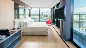 A modern hotel room with a large bed, a TV, a red chair, and a city view. - Barceló Hamburg (Hamburg)
