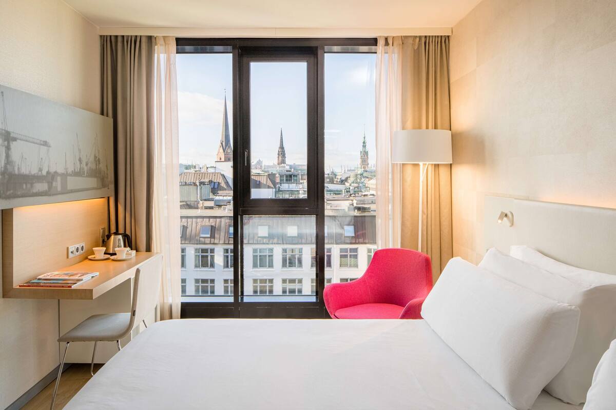 A hotel room with a large bed, a desk with a chair, a red chair, and a view of the city through a large window.
