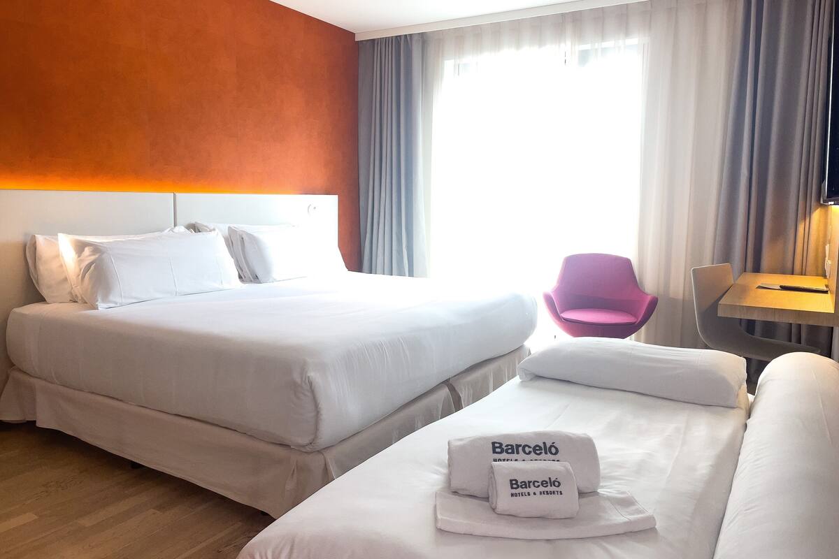 A hotel room with two beds, a desk with a chair, and a window with curtains.
