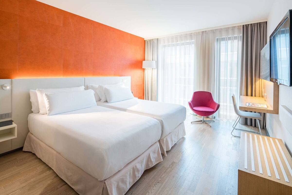 A hotel room with two beds, a desk, a chair, a TV, and a window with curtains.