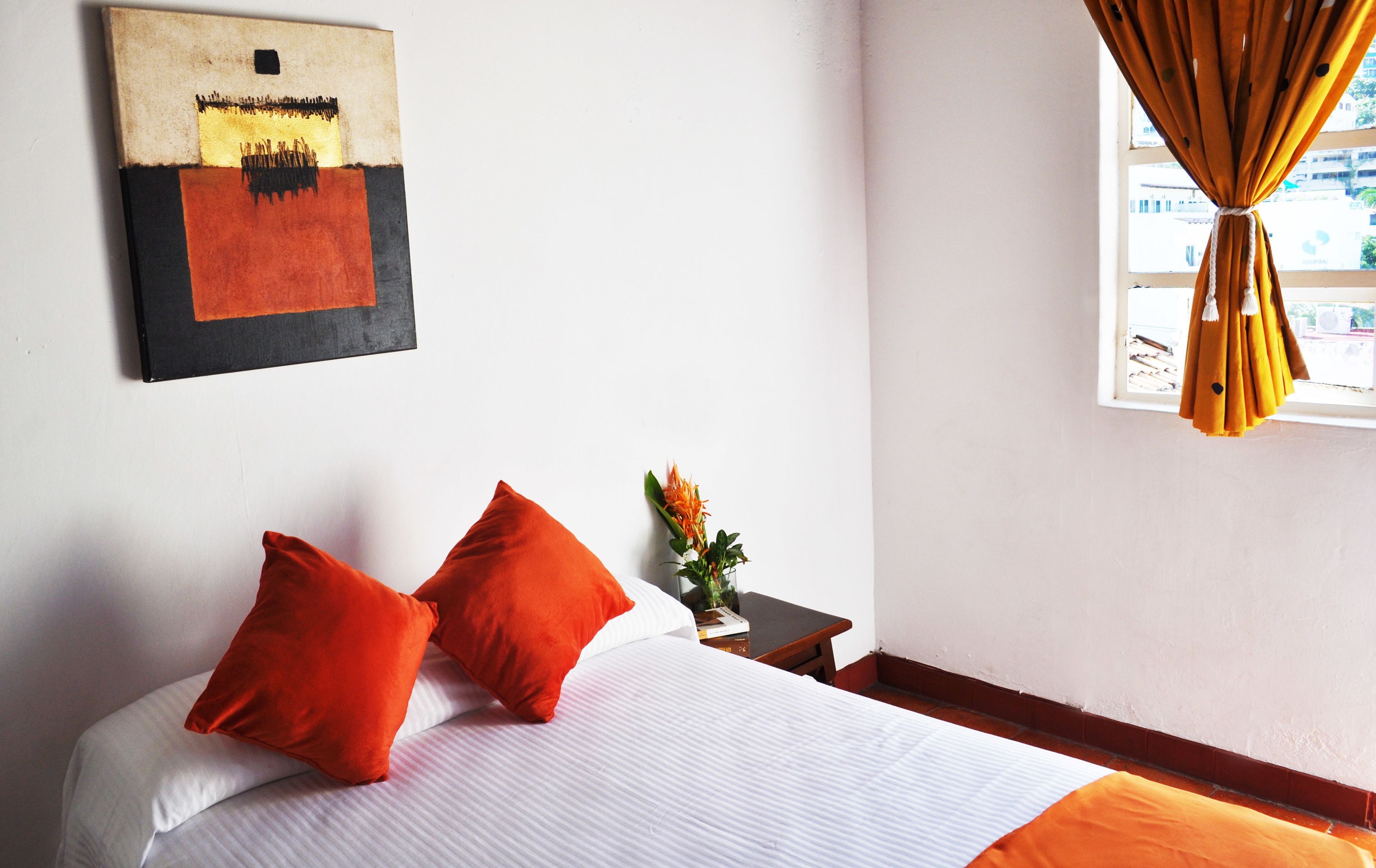A hotel room with a bed, two orange pillows, a small table with a plant, and a painting on the wall.