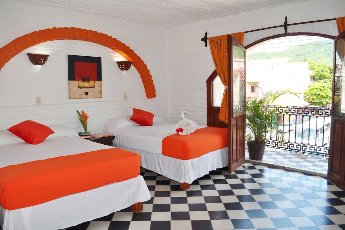 Habitación de hotel con dos camas, suelo a cuadros, balcón con vistas y pared de ladrillo blanco.