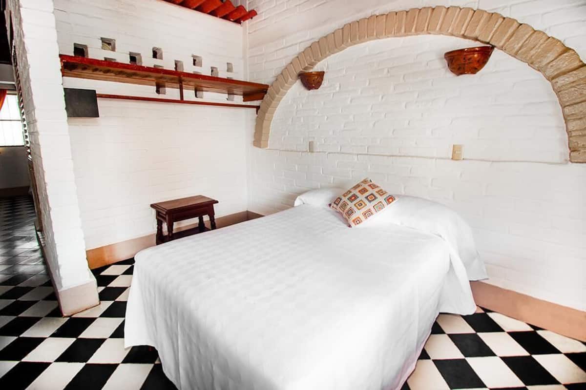 Una habitación con piso a cuadros, una cama con ropa blanca, una pequeña mesita de noche de madera y un arco de ladrillo.