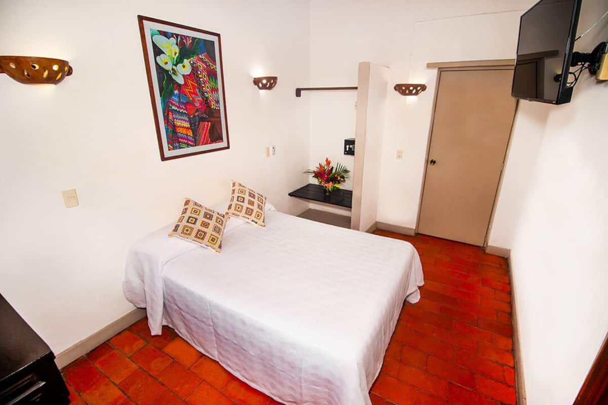 Una habitación de hotel con cama, mesita de noche, televisor, un cuadro y una puerta.