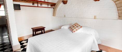 A room with a checkered floor, a bed with white bedding, a small wooden nightstand, a white brick wall, and a decorative archway.