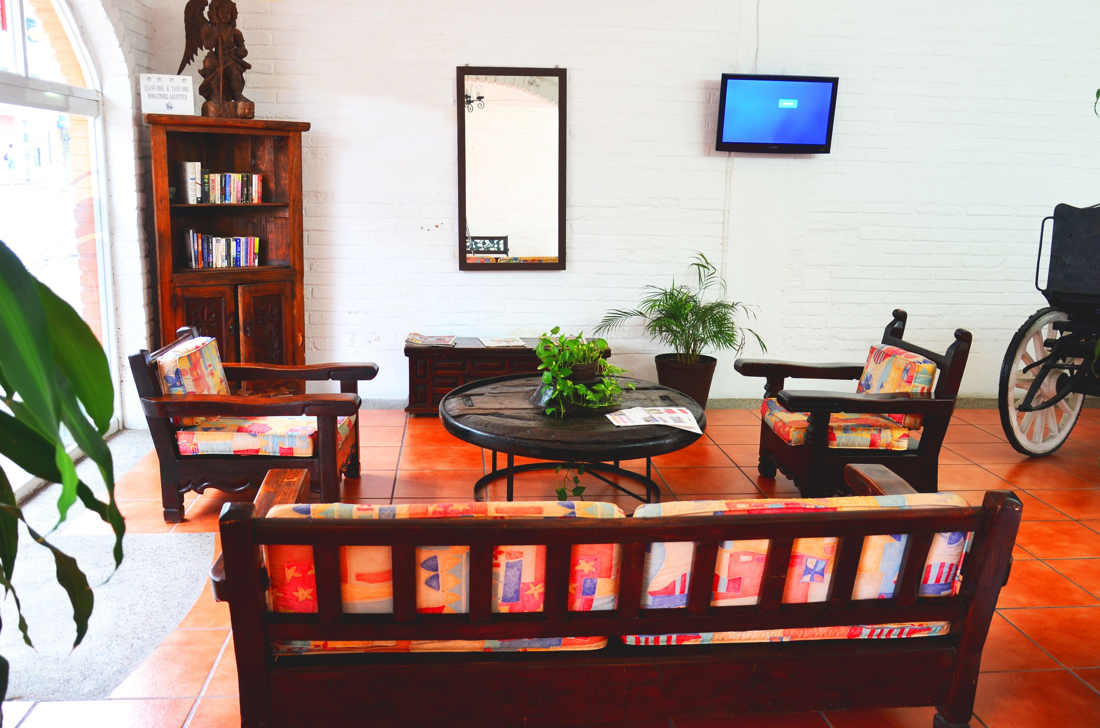 A living room with a sofa, chairs, a table, a bookshelf, a potted plant, a wheelchair, and a TV on the wall.