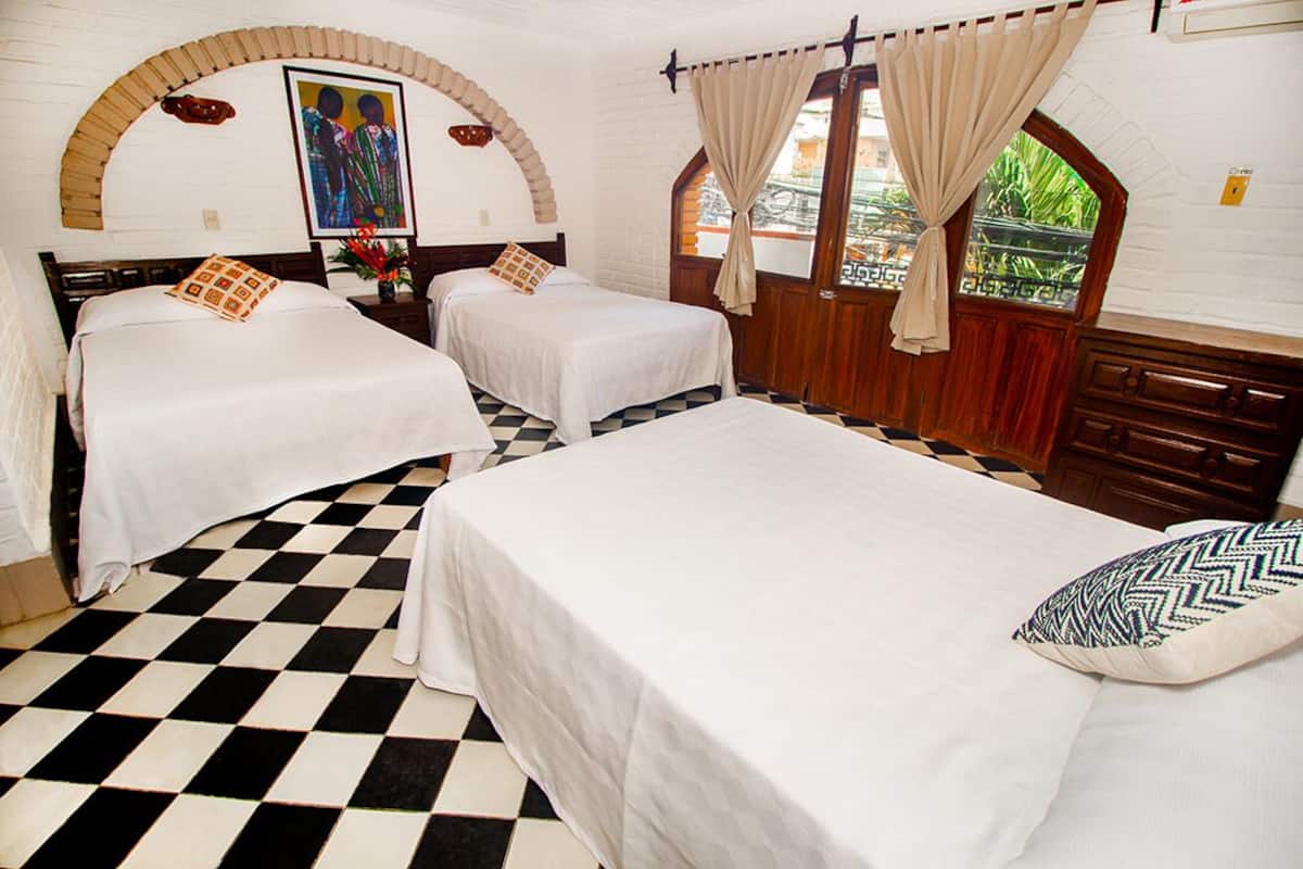 Habitación de hotel con tres camas, suelo a cuadros, ventana en arco y un cuadro en la pared.
