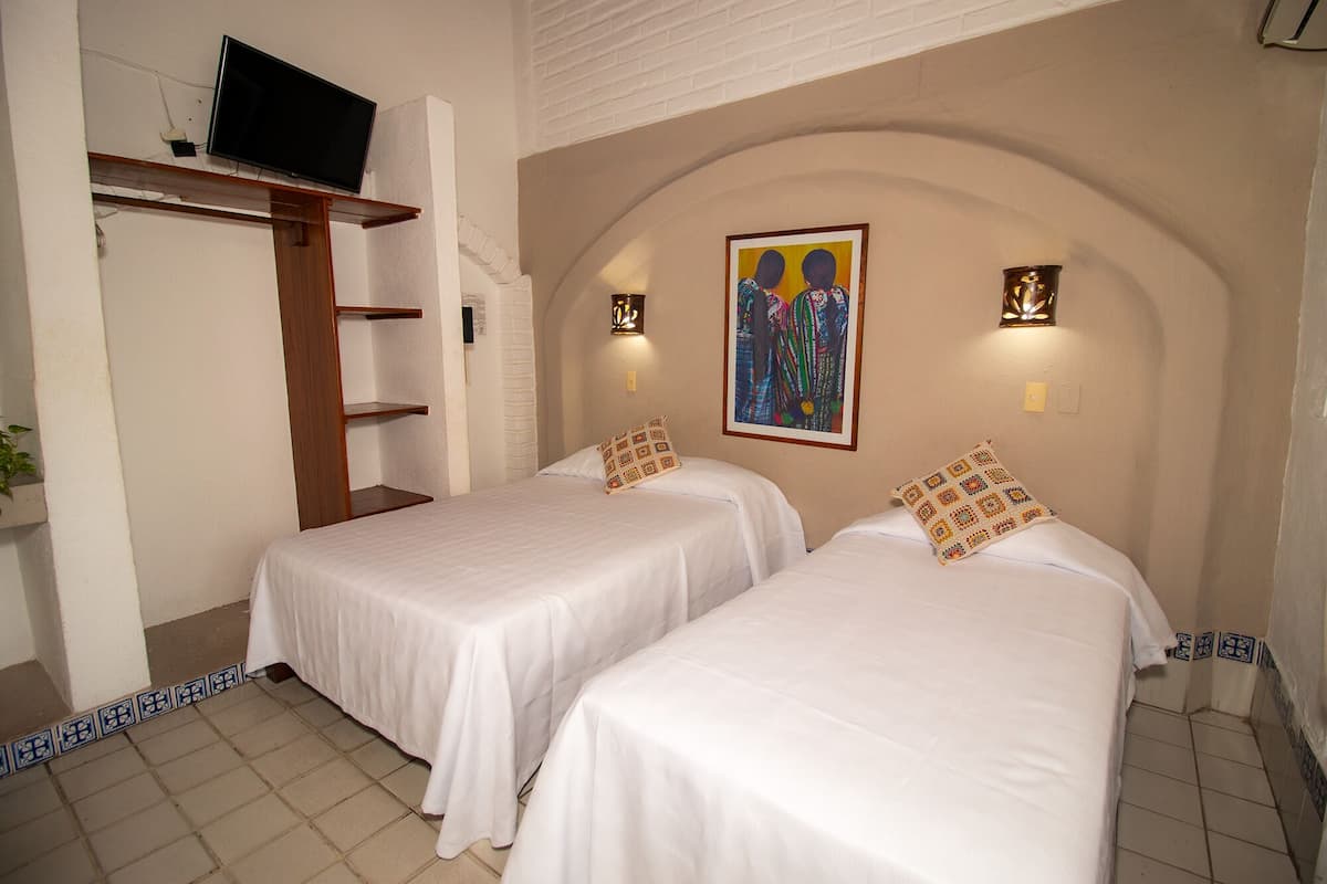 Habitación de hotel con dos camas individuales, un televisor montado en una repisa, un cuadro en la pared y una planta pequeña a la izquierda.