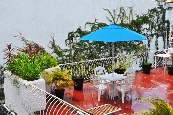 Una terraza en la azotea con una mesa y sillas blancas, un parasol azul y diversas macetas con plantas.