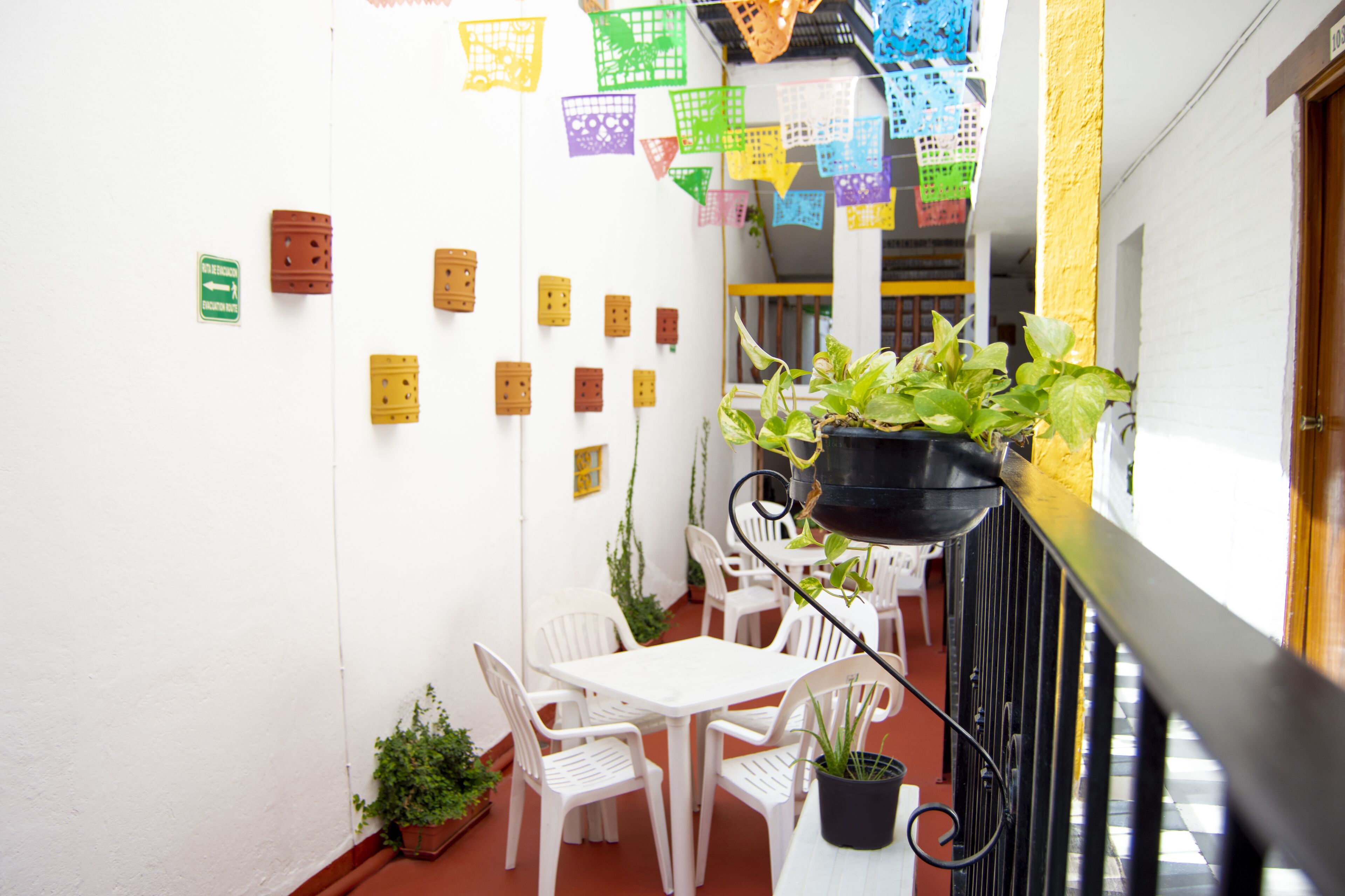 Un pequeño patio al aire libre con sillas y mesas de plástico blanco, una planta en maceta y decoraciones de papel de colores en la pared.