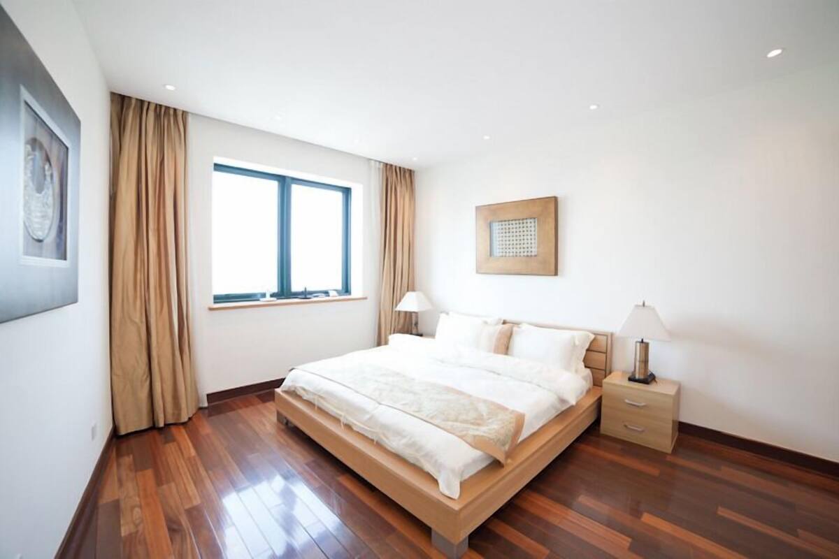 Ein modernes Schlafzimmer mit einem großen Bett, zwei Nachttischen, einem Fenster mit Vorhängen und einem gerahmten Bild an der Wand.