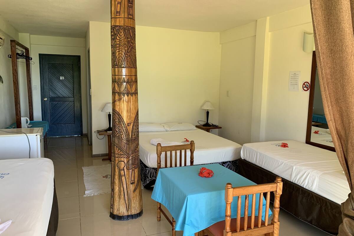 A hotel room with two single beds, a wooden chair, a table with a blue tablecloth, a wooden pillar with carvings, a door, and a mirror.