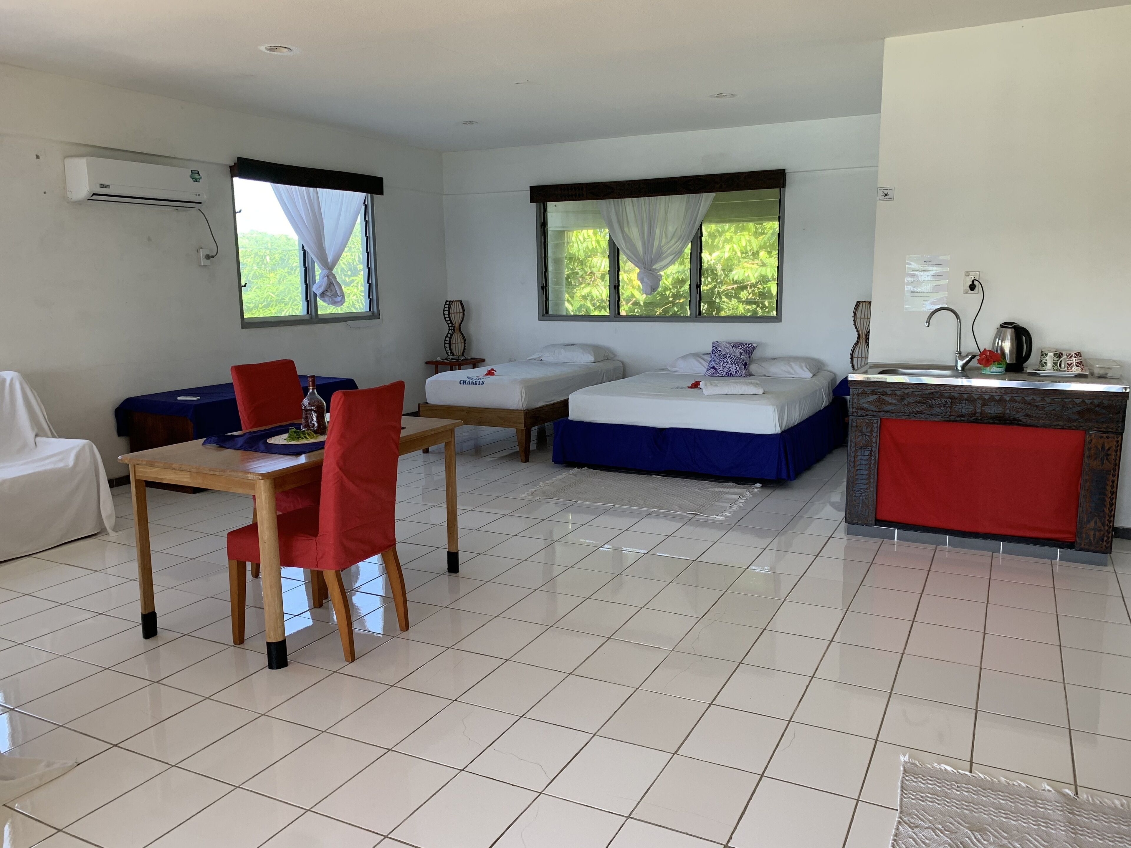 A hotel room with a bed, a small table with chairs, a desk, a window with curtains, and a kitchenette.