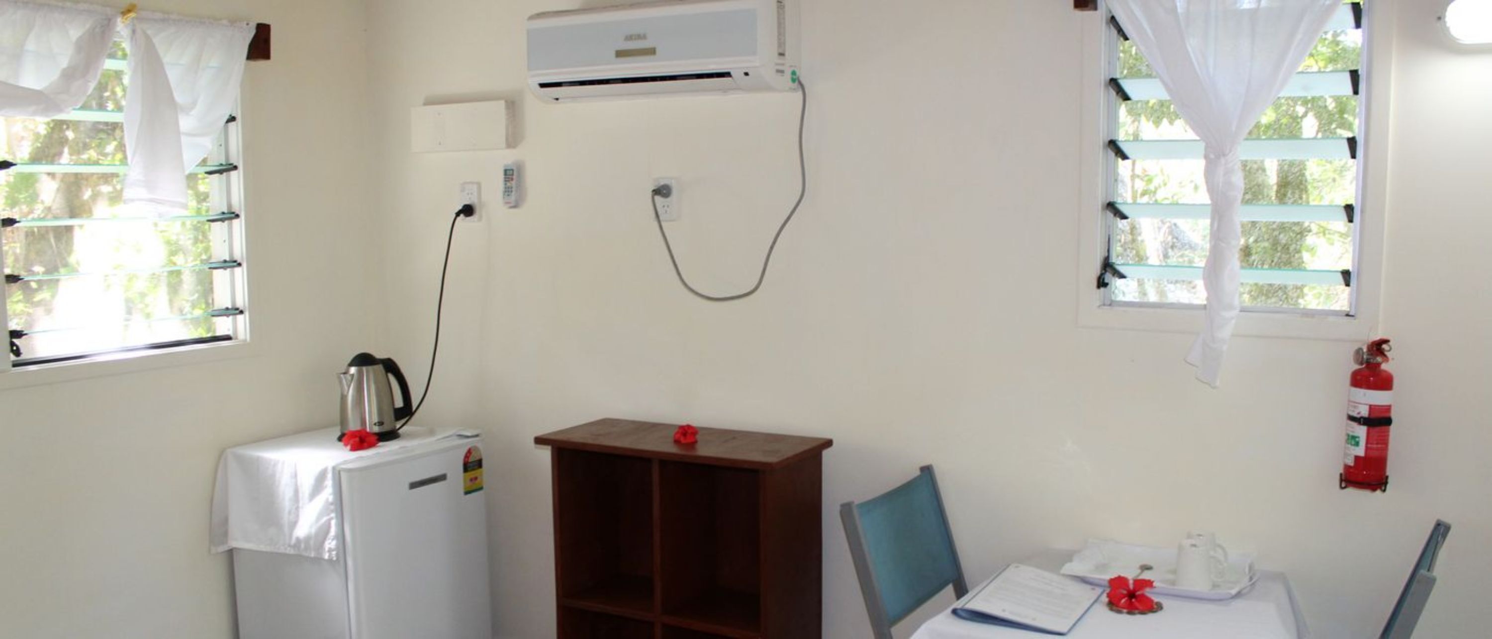 Una habitación pequeña y bien iluminada con un refrigerador blanco, un armario de madera, una mesa con una silla y una ventana con cortina blanca.