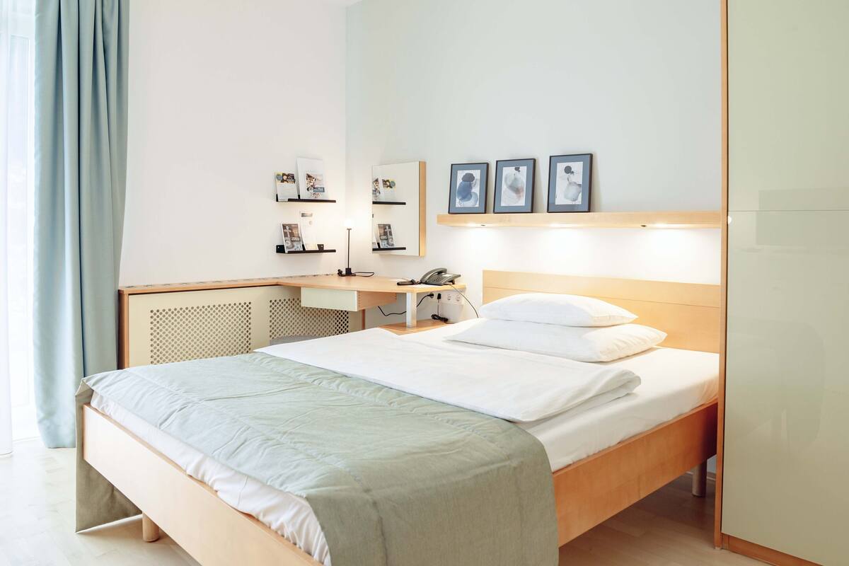 A neatly arranged hotel room with a bed, bedside table, lamp, and wall-mounted shelves with framed pictures.