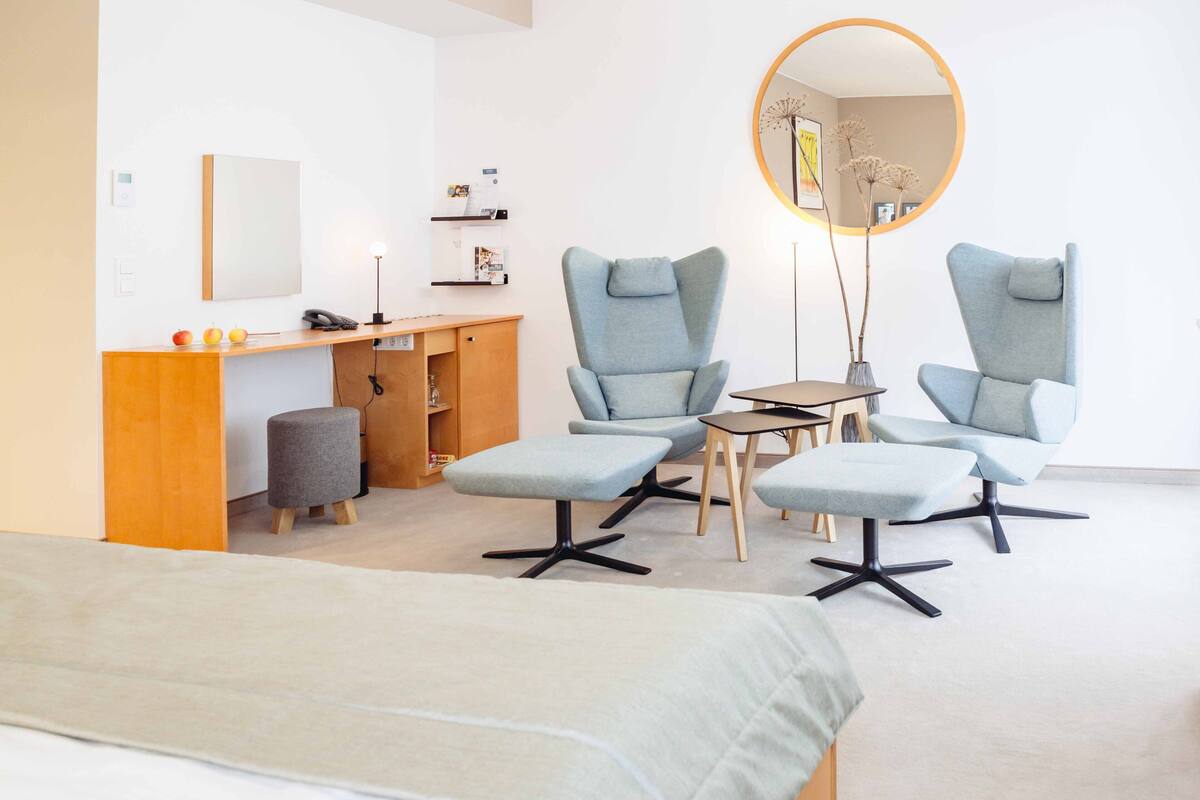 A modern room with a bed, two armchairs, a small table, a desk, and a mirror.