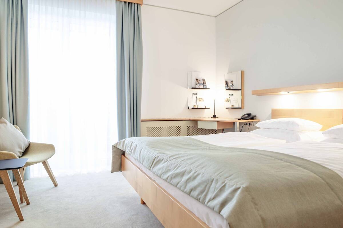 A hotel room with a bed, a desk, a chair, and a large window with curtains.