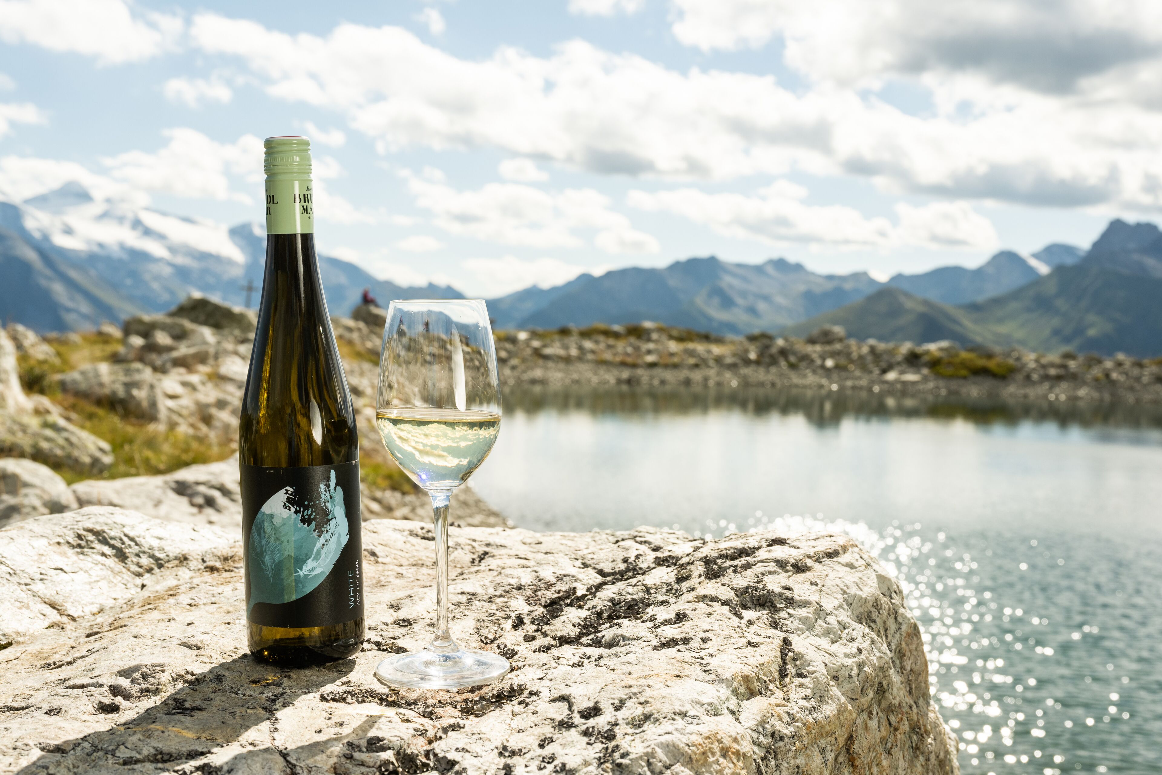 Uma garrafa de vinho e um copo sobre uma superfície rochosa, com um lago e montanhas ao fundo.