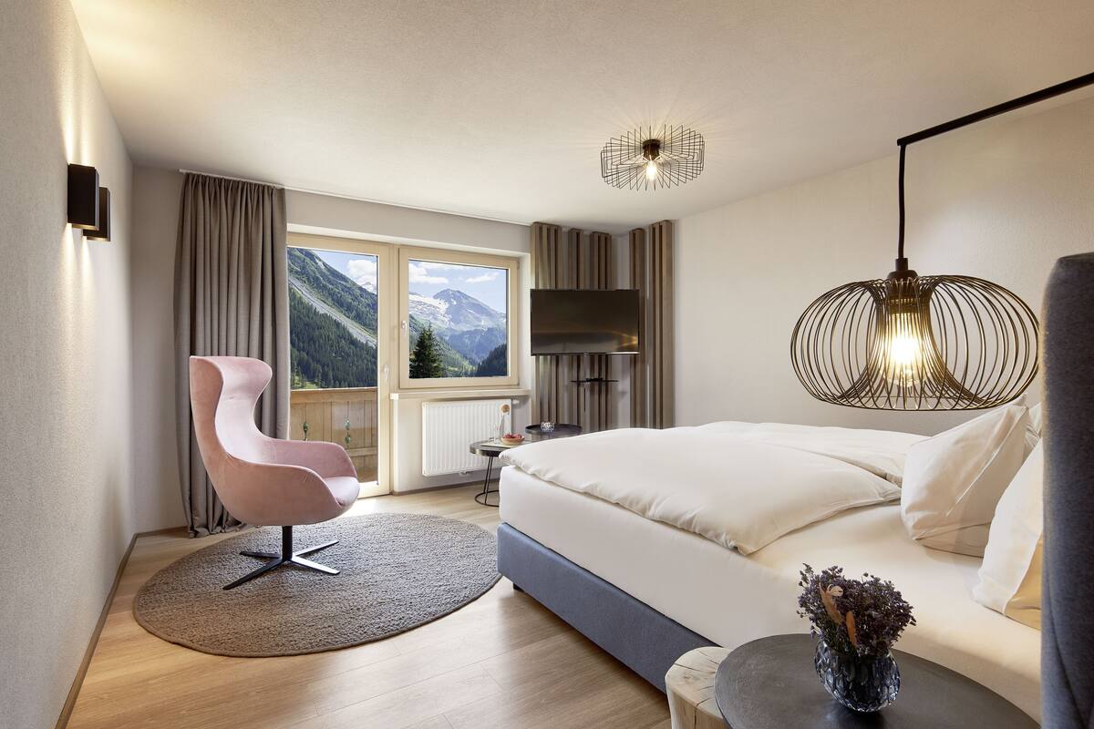 Un hotel moderno con una cama grande, una butaca curvada, televisión y vistas a montañas desde la ventana.