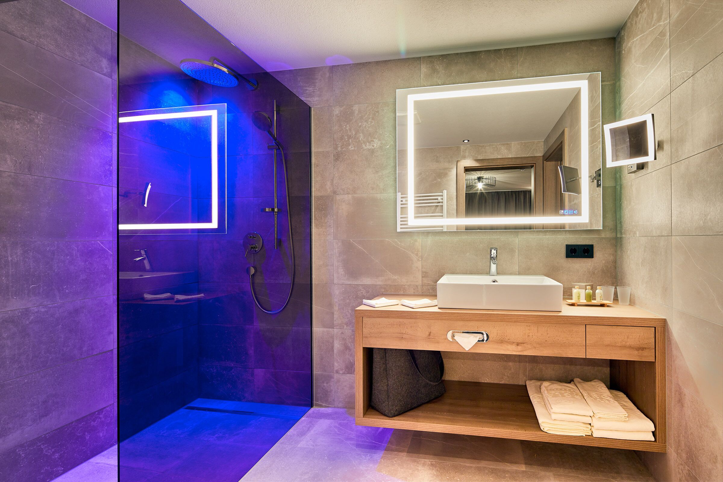 Un baño moderno con ducha de vidrio, tocador de madera con espejo y toallero empotrado.