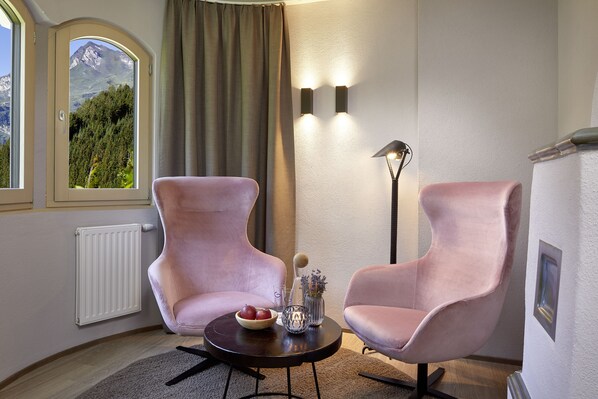 A room with two pink armchairs, a small round table with a bowl of fruit, a vase with flowers, and a floor lamp. A window with a mountain view is visible. - Adler Inn Tyrol Mountain Resort SUPERIOR (Tux)