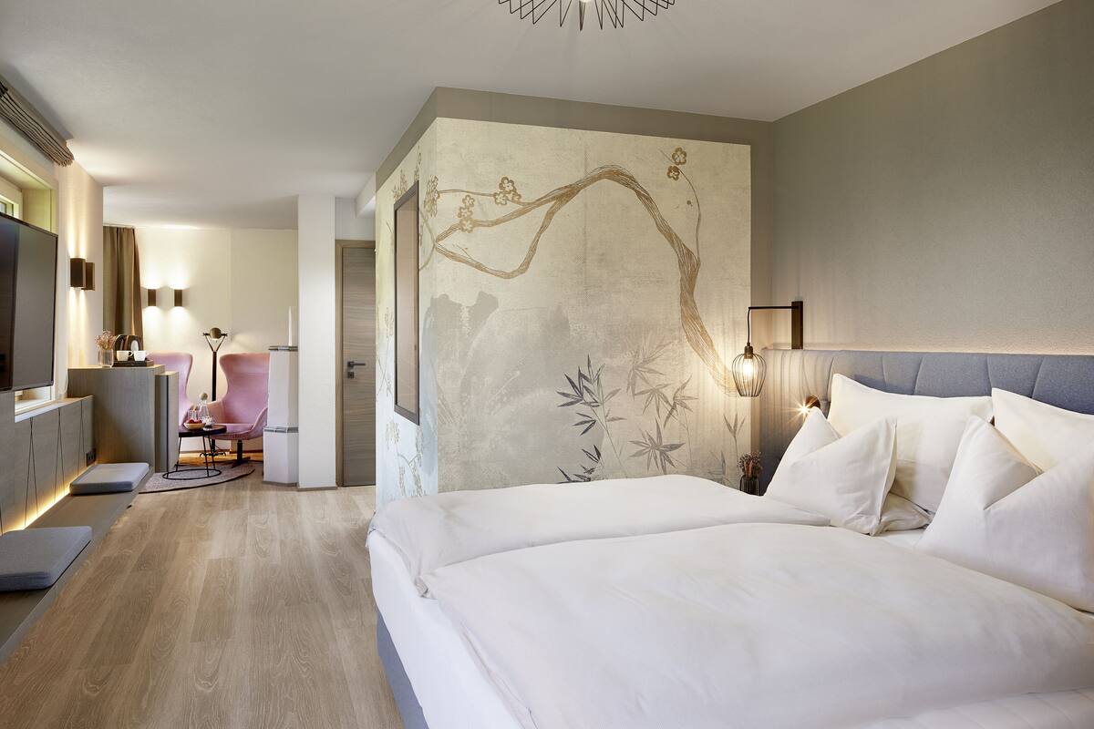 Una habitación de hotel moderna con una cama grande, una zona de estar con una silla y una mesa, y una pared decorativa con motivos florales.
