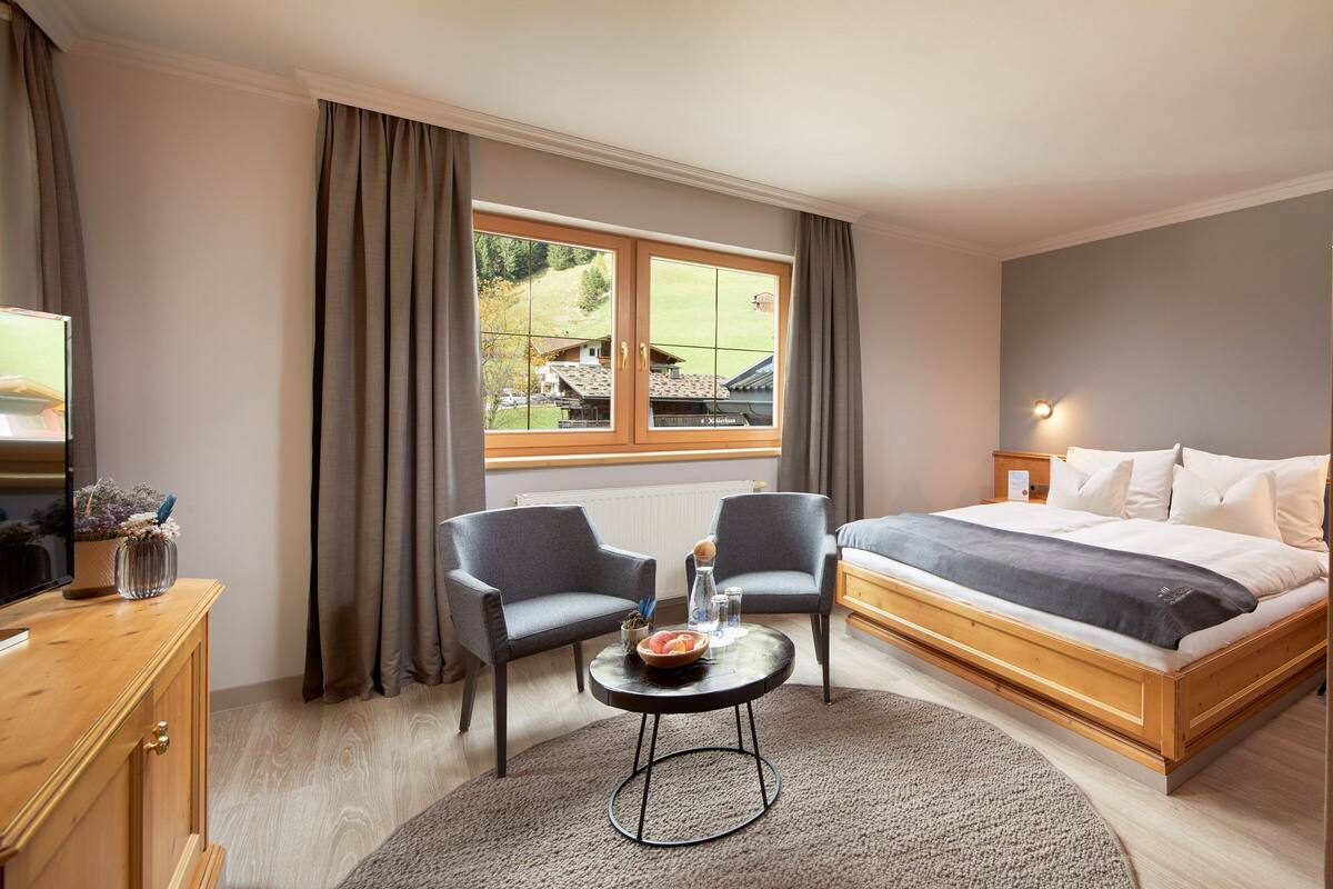 Un dormitorio con cama, dos sillas, una mesita, televisión y ventana con cortinas.