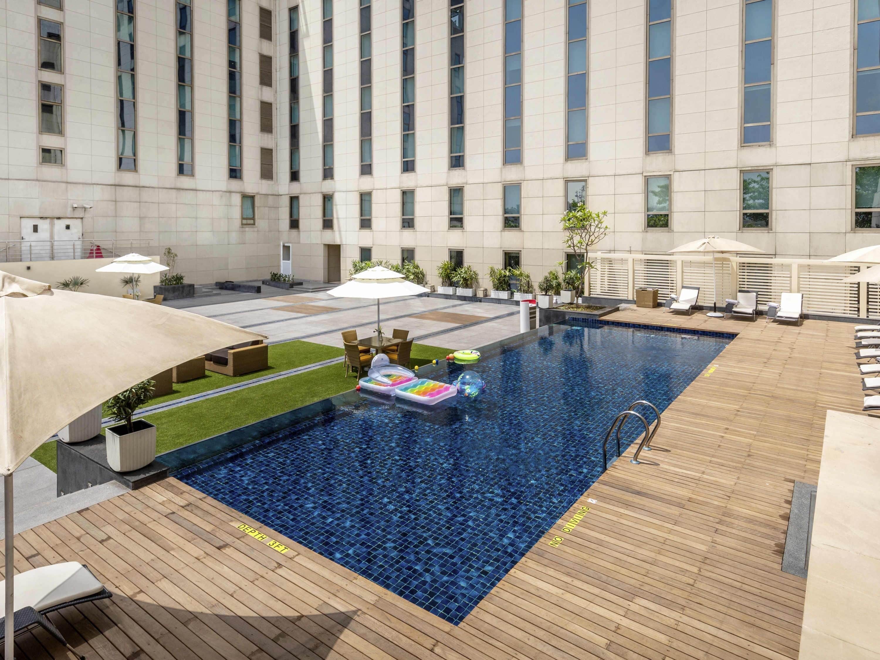 A rooftop pool with a depth marker, lounge chairs, umbrellas, and a view of a tall building.