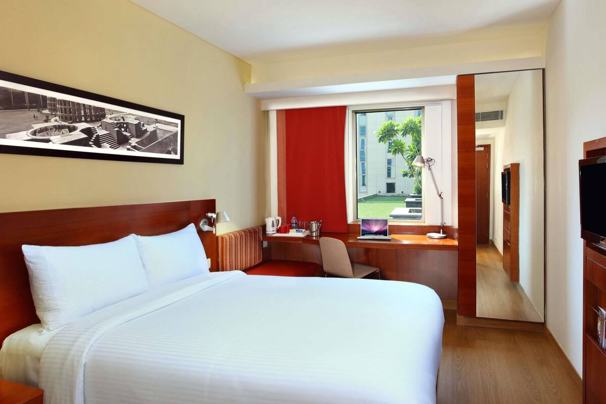 Ein Hotelzimmer mit einem großen Bett, einem Schreibtisch mit einem Laptop, einem Fernseher und einem Fenster mit Blick auf ein Gebäude und Grünflächen.