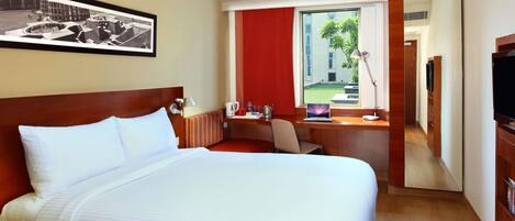 A hotel room with a large bed, a desk with a laptop, a TV, and a window with a view of a building and greenery.