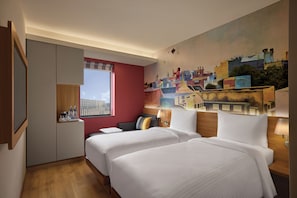 Une chambre d’hôtel avec deux lits, une fresque représentant un paysage urbain, une fenêtre avec un cadre rouge et un plancher en bois.