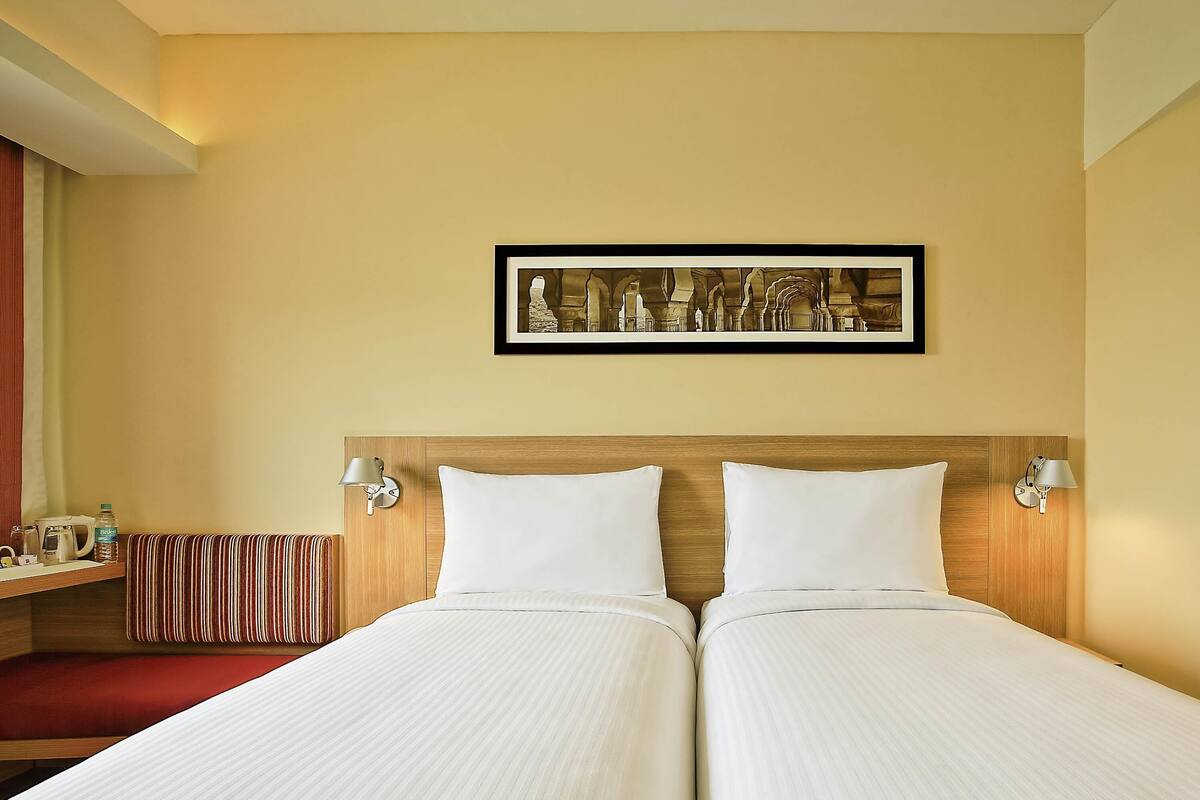 Ein Hotelzimmer mit zwei Betten, einem Holzkopfteil, Nachttischlampen, einem gerahmten Bild an der Wand und einer roten Bank.