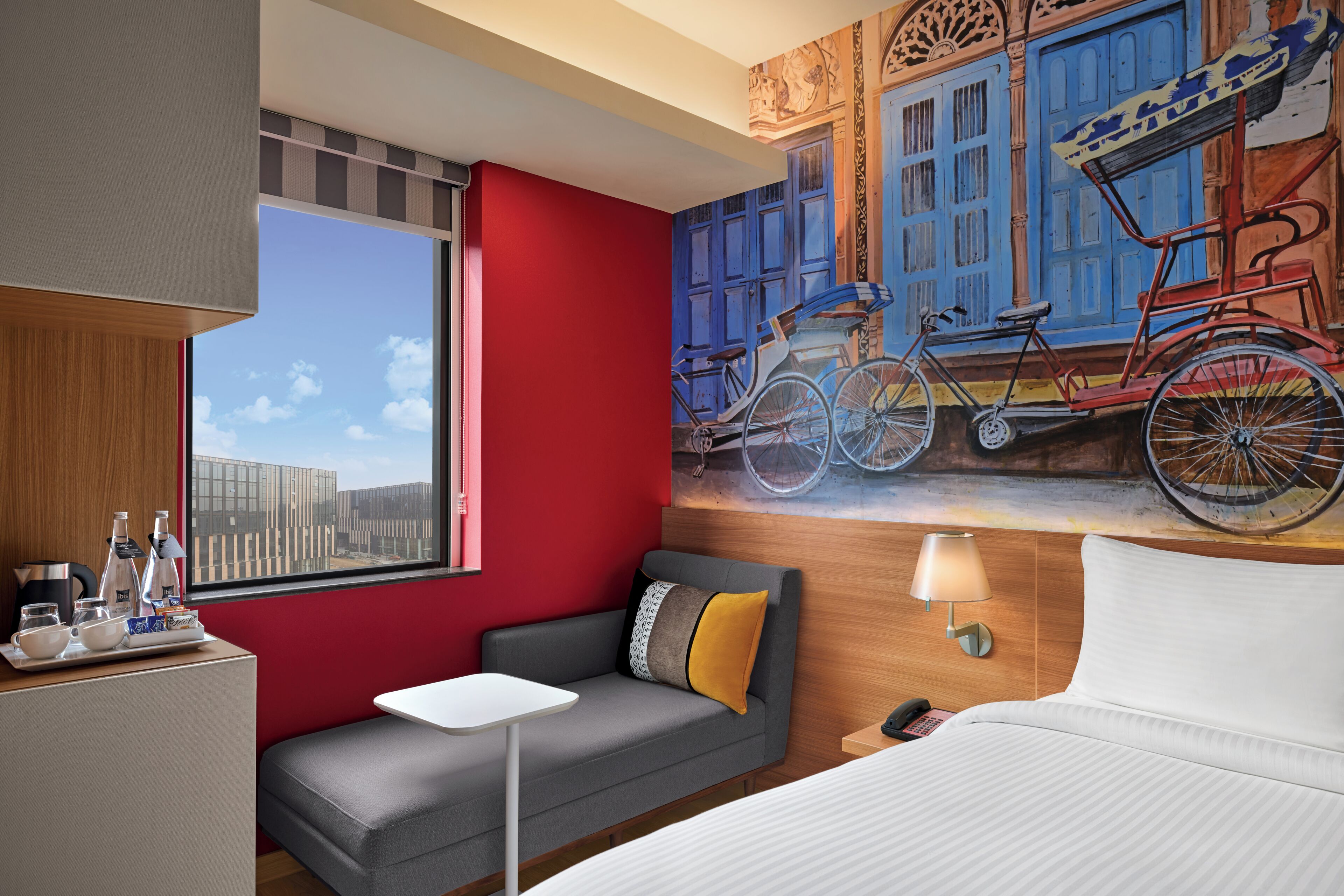 A hotel room with a red wall, a gray sofa, a white bed, a small table, a lamp, and a mural of bicycles.