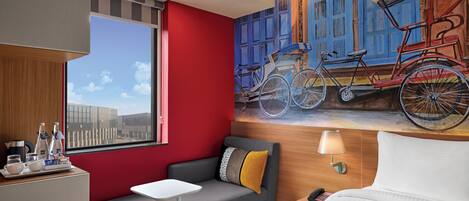 A hotel room with a red wall, a gray sofa, a white bed, a small table, a lamp, and a mural of bicycles.