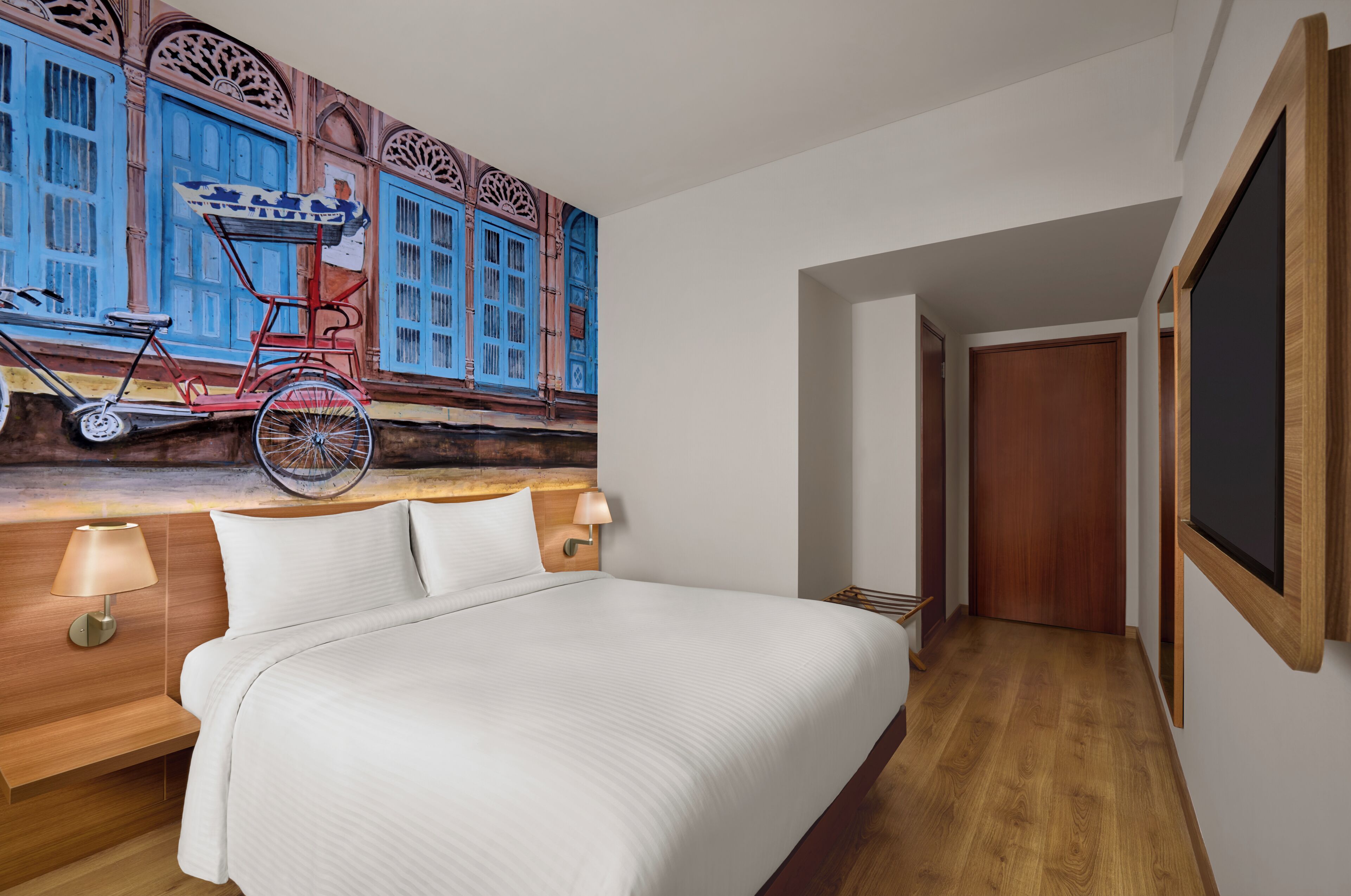 A hotel room with a bed, a wall-mounted TV, a wooden nightstand with a lamp, and a mural of a bicycle.