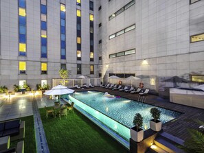 A rooftop pool area with lounge chairs and umbrellas, surrounded by a high-rise building with illuminated windows.