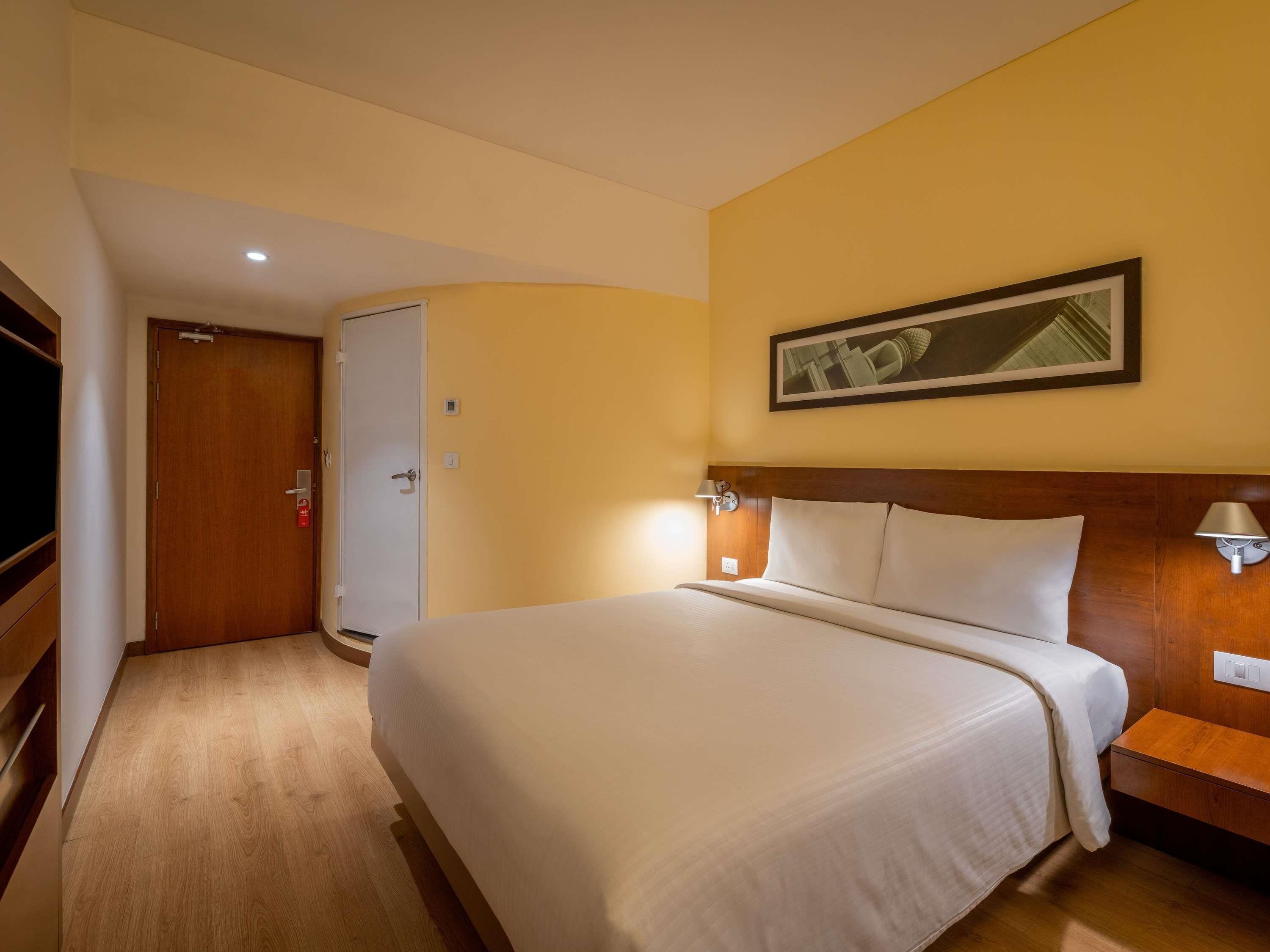 A hotel room with a bed, a wooden headboard, a nightstand, a wall-mounted lamp, a door, and a framed picture on the wall.