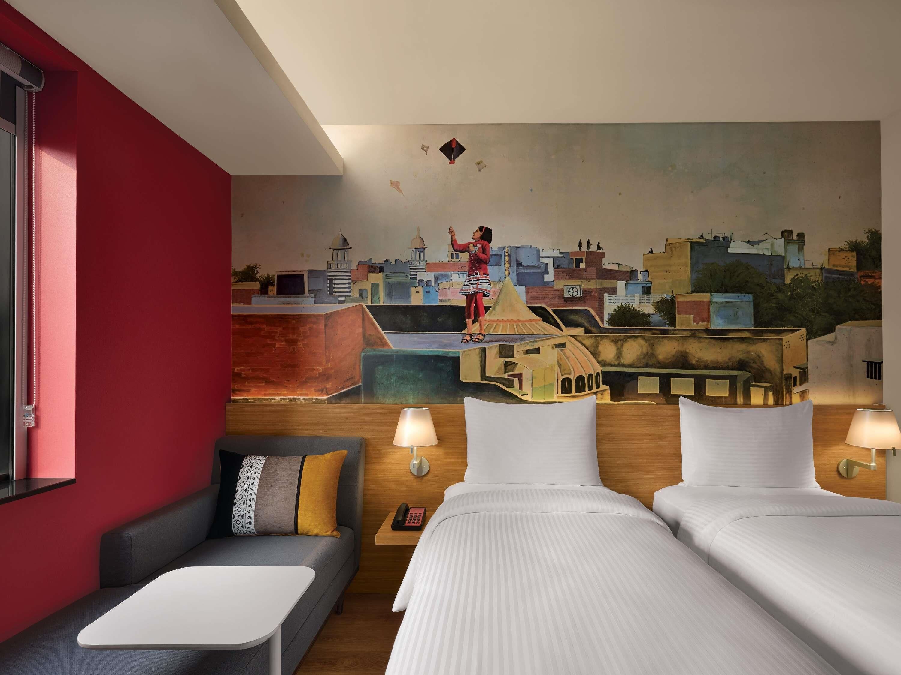 Une chambre d’hôtel avec une fresque représentant un paysage urbain, deux lits, un canapé et une petite table.