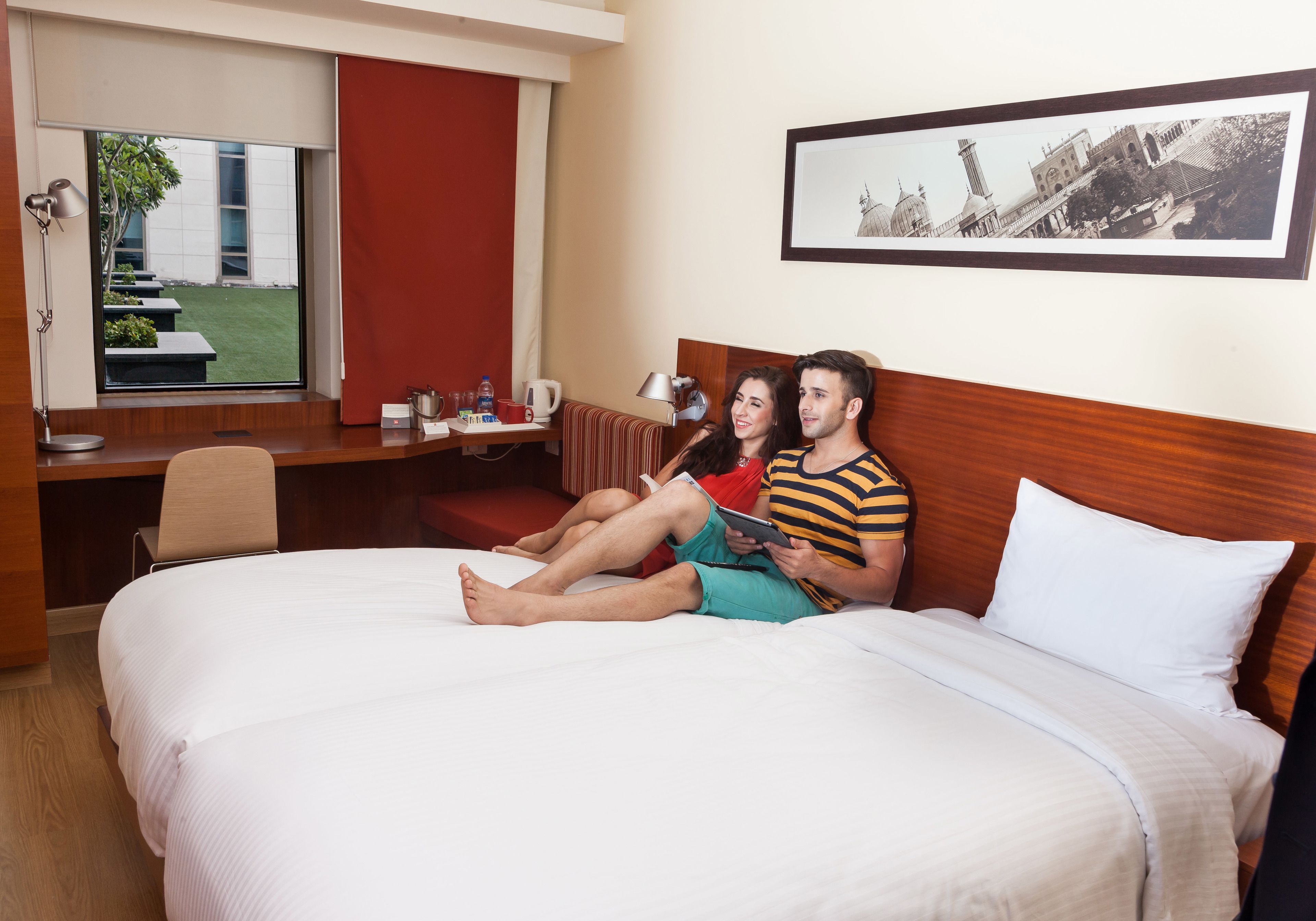 A hotel room with a large bed, a desk with a chair, a window with a view of a building, and a framed picture on the wall.